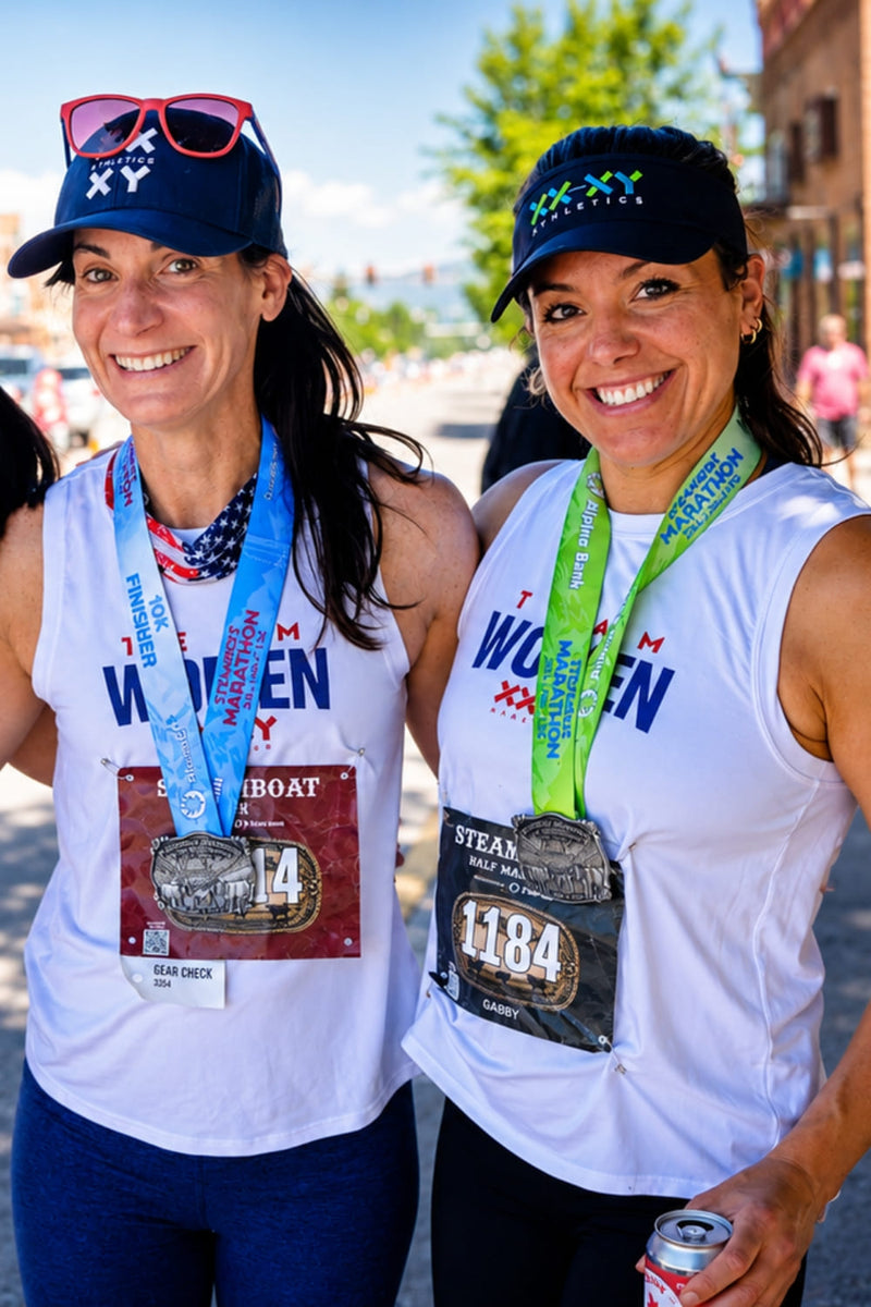 Two smiling women wearing white tank tops with 'WOMEN' printed on them, race bibs, and colorful event lanyards, standing outdoors after a race. One wears a blue cap with 'XXY' and sunglasses on top, and the other wears a dark cap with multicolor lettering.