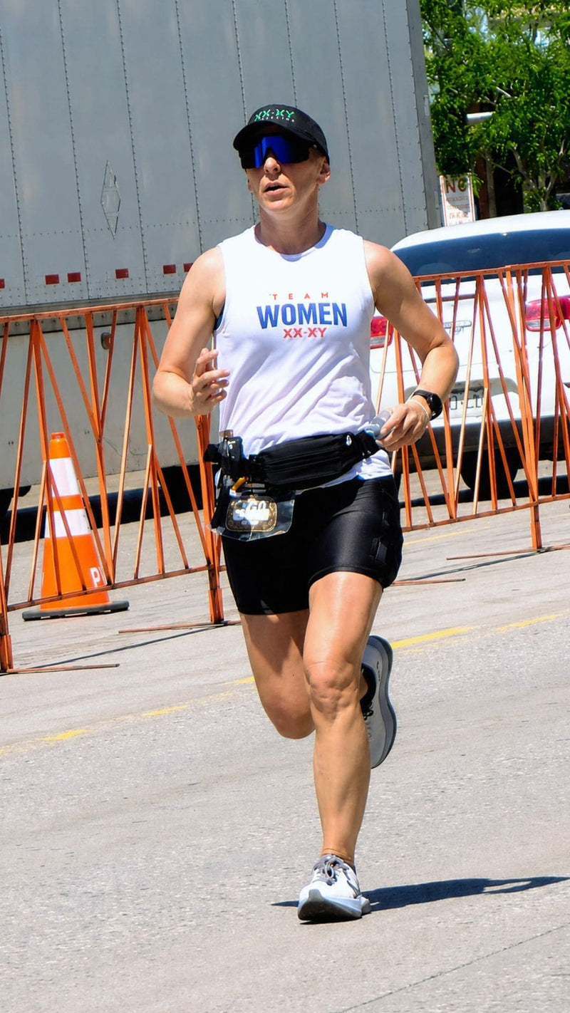 A woman running outdoors wearing a white tank top with 'RUN WOMEN 5K 20K' printed on the front, black athletic shorts, a black waist pack, and a black cap. She is running on a paved road with orange barricades and a large truck in the background.