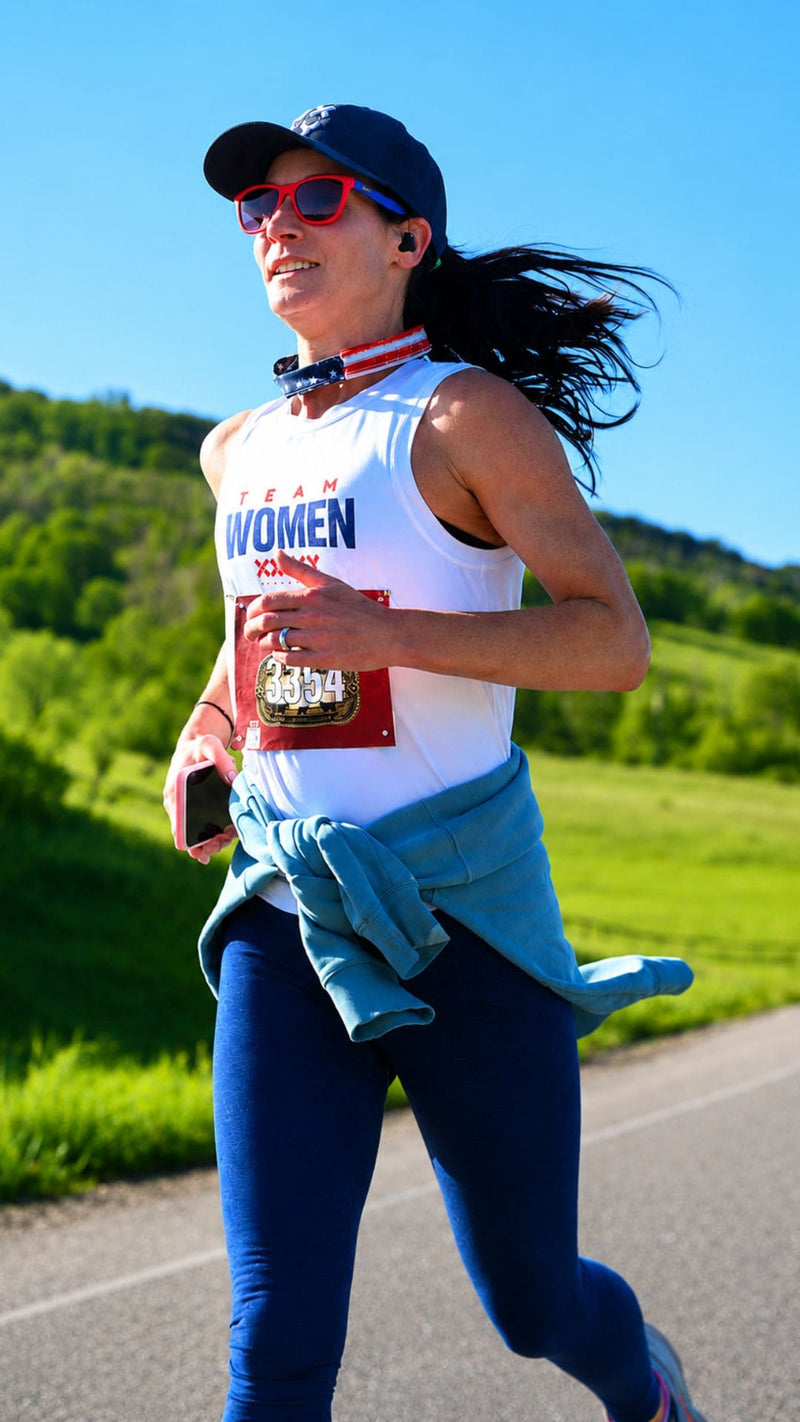 A woman running outdoors on a road wearing a white sleeveless athletic top with 'TEAM WOMEN' and a race bib, blue leggings, a blue sweatshirt tied around her waist, a dark cap, and red sunglasses.
