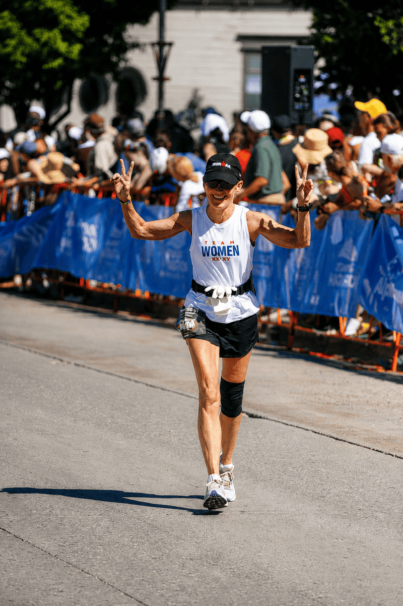 A female marathon runner crosses the finish line on a city street, wearing a white tank top with 'WOMEN' printed in blue, black shorts, and a black cap. She is smiling and making peace signs, with a crowd and blue race barriers in the background.