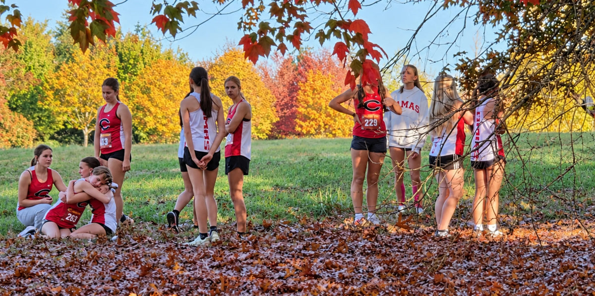 Camas High School girls’ cross-country runners gather beneath colorful autumn trees after a race, with two teammates embracing while others stand nearby in red and white uniforms.