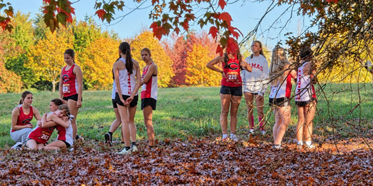 Camas High School girls’ cross-country runners gather beneath colorful autumn trees after a race, with two teammates embracing while others stand nearby in red and white uniforms.