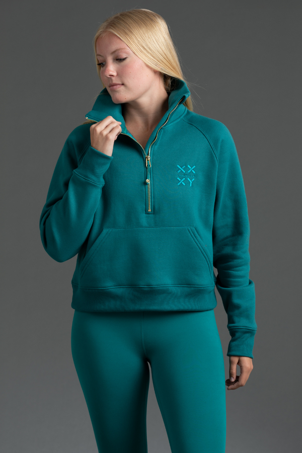 Person wearing a teal tracksuit with a logo on a gray background || Teal