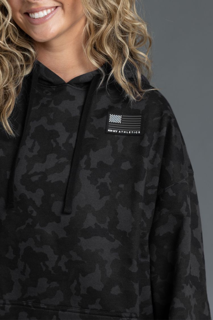 Person wearing a black camouflage hoodie with a logo on a gray background
