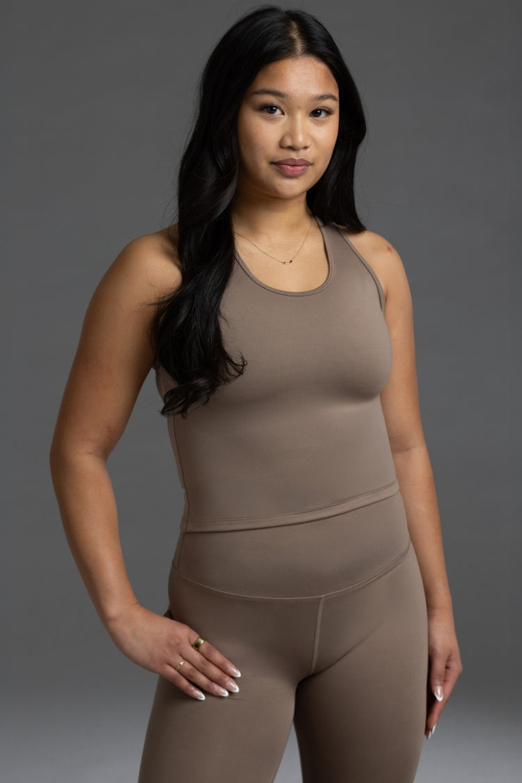 A woman modeling a matching two-piece shapewear set in a solid taupe-brown color, consisting of a sleeveless tank top and high-waisted leggings, against a neutral gray studio background. || Mocha