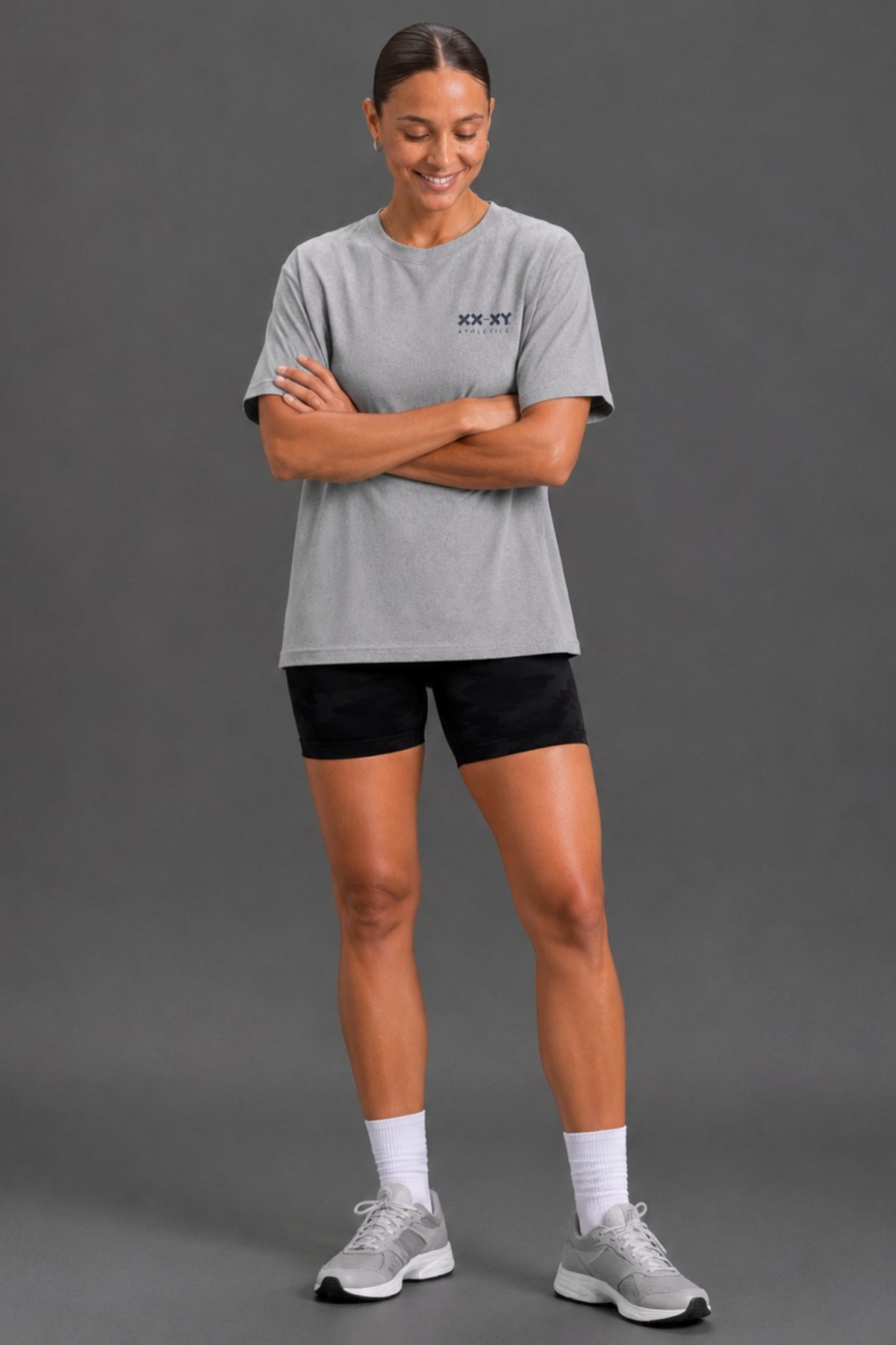 A woman modeling a light gray heathered short-sleeve t-shirt with a small black 'XX-SKY' logo on the left chest, paired with black athletic shorts, white socks, and gray running shoes, against a solid dark gray studio background.