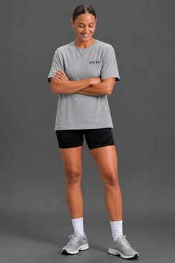 A woman modeling a light gray heathered short-sleeve t-shirt with a small black 'XX-SKY' logo on the left chest, paired with black athletic shorts, white socks, and gray running shoes, against a solid dark gray studio background. || Heather Gray