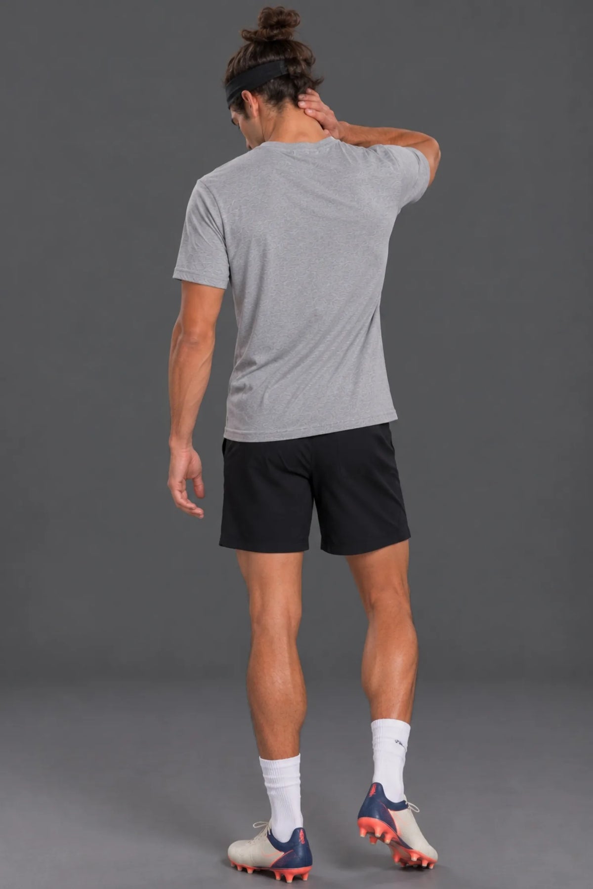 Male model wearing a heather gray XX-XY Athletics USA Flag V-Neck Tee, black athletic shorts, and soccer cleats, shown from behind against a gray studio background.