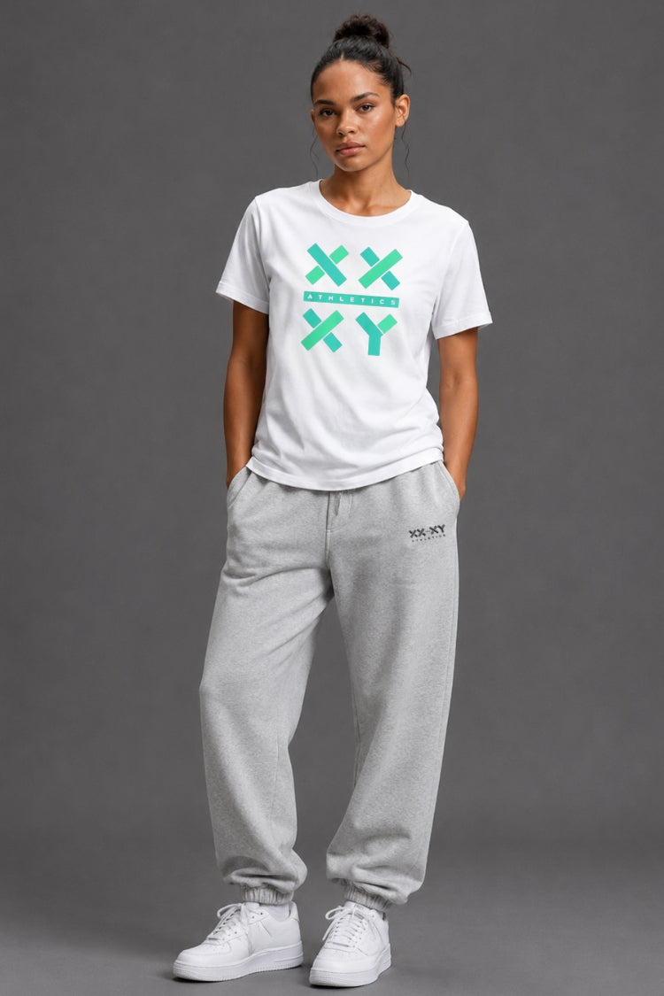 A woman modeling a white crewneck t-shirt with a green XXYY graphic and light gray sweatpants, standing against a solid gray studio background.