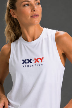 A woman wearing a white sleeveless athletic tank top with a navy and red 'XX-XY ATHLETICS' logo on the chest, photographed against a dark gray studio background. || White