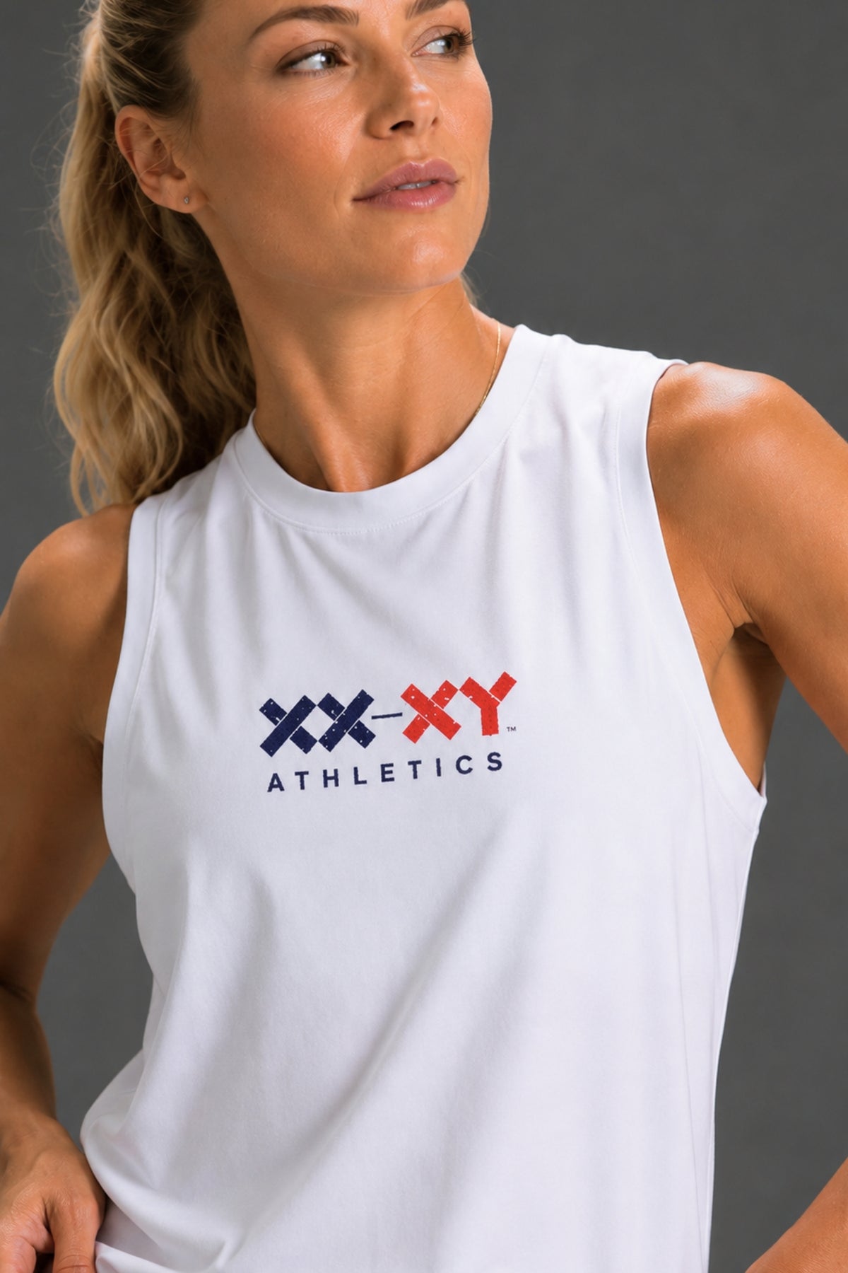 A woman wearing a white sleeveless athletic tank top with a navy and red 'XX-XY ATHLETICS' logo on the chest, photographed against a dark gray studio background. || White