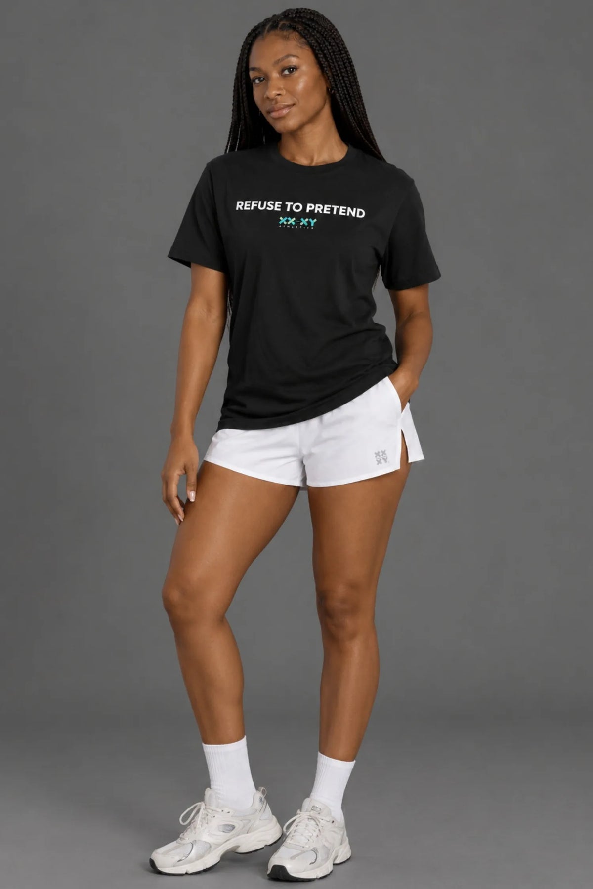 Female model standing in black Refuse to Pretend graphic tee, white athletic shorts, and sneakers against gray studio background. || Black