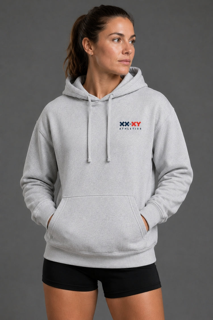 A woman wearing a light gray pullover hoodie with a small red and blue 'XX-XY Athletics' logo on the left chest, paired with black athletic shorts, against a solid dark gray studio background. || Heather Gray