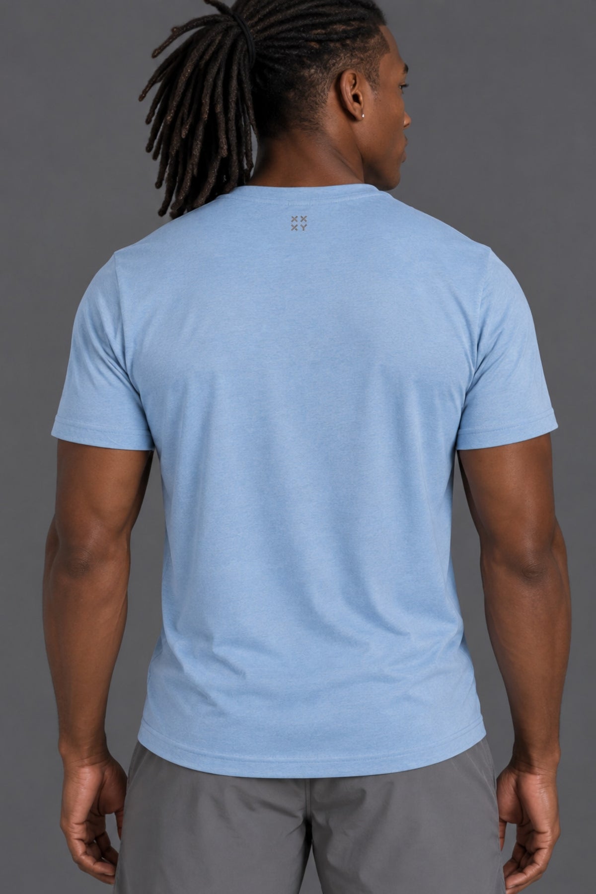 A man with dreadlocks is shown from the back wearing a light blue crewneck t-shirt and gray shorts against a solid gray studio background. || Cerulean Blue Heather