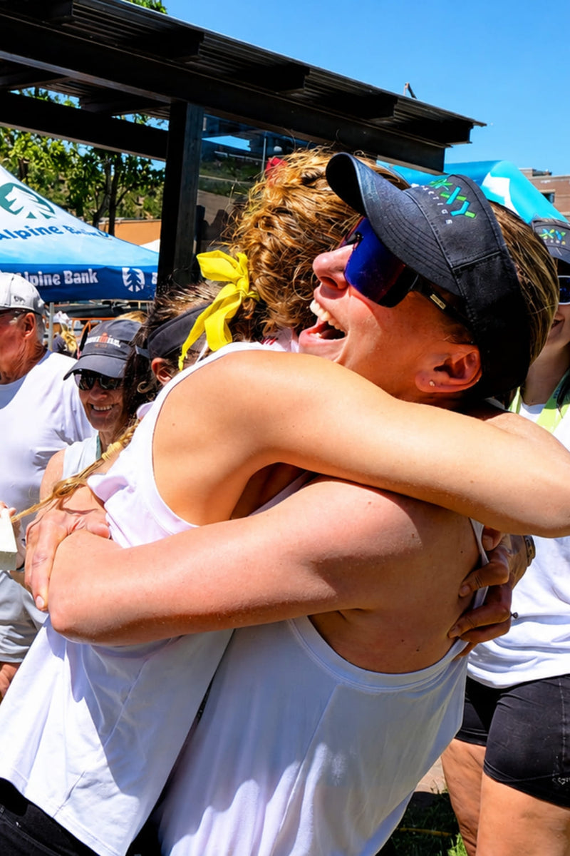 Two women hugging outdoors under a covered walkway. One wears a white tank top and black visor, and the other is mostly obscured but wearing a yellow hair tie.