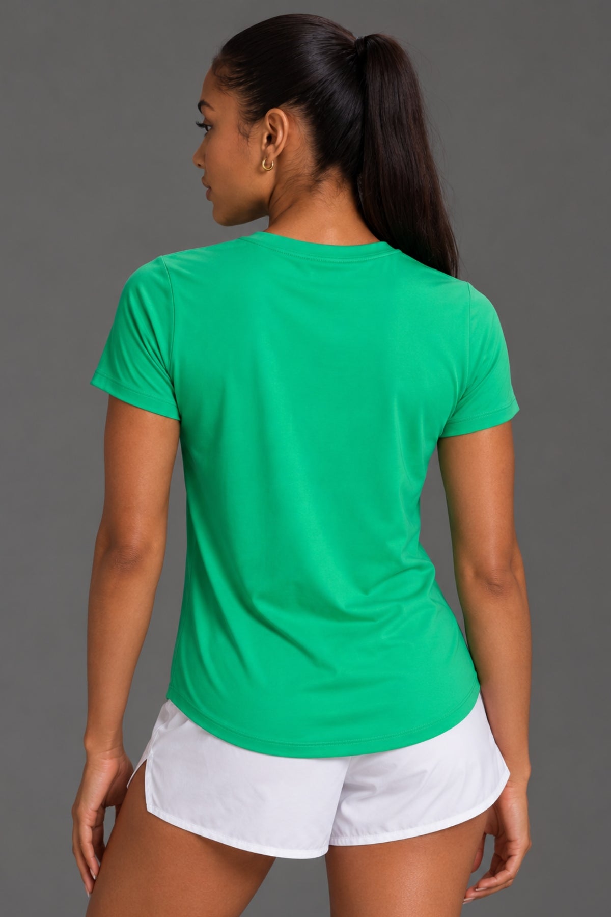 A woman shown from the back wearing a bright green short-sleeve athletic T-shirt and white shorts against a plain gray studio background. || Kelly Green