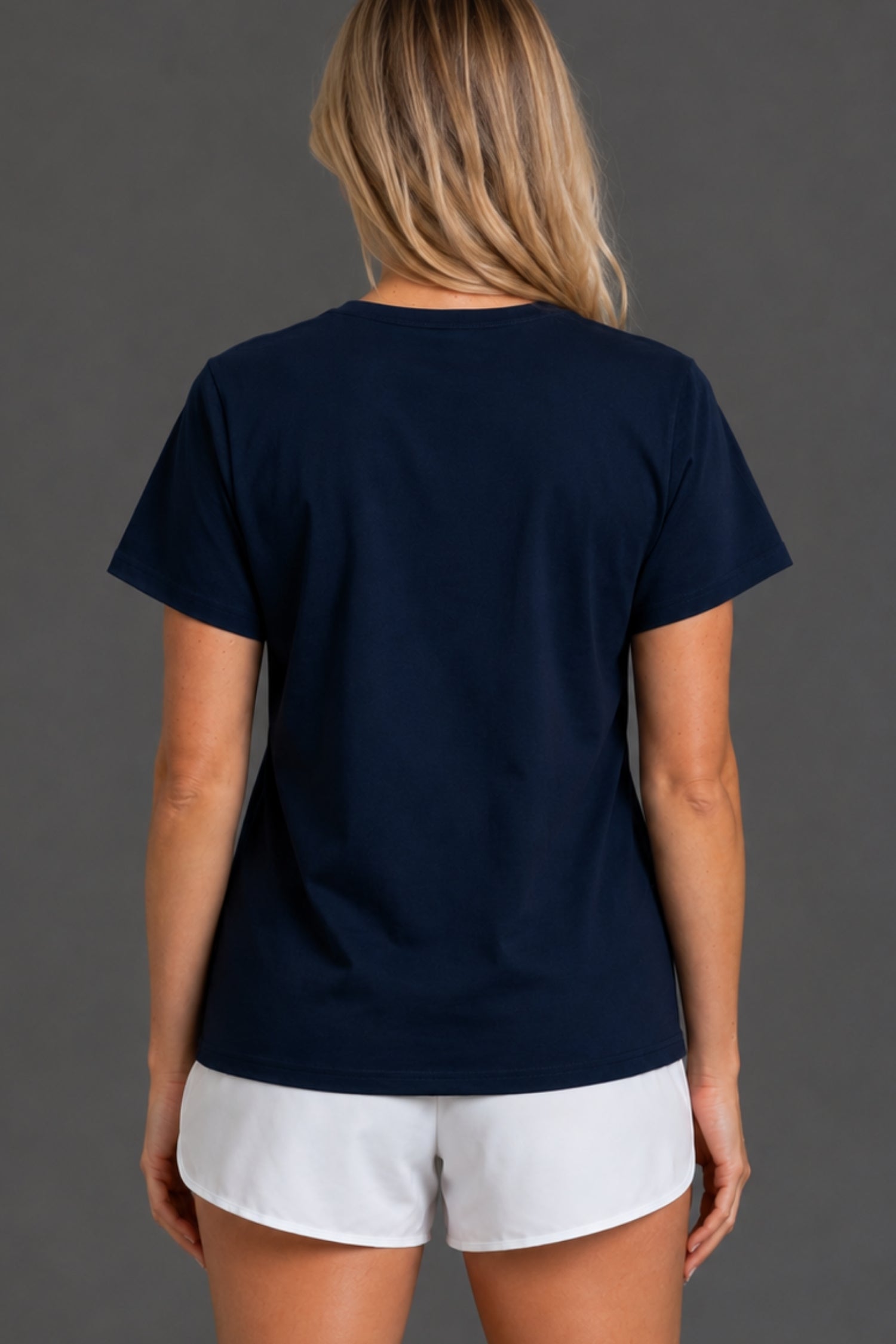 A woman viewed from the back wearing a navy blue short-sleeve crewneck t-shirt and white shorts, set against a solid dark gray studio background. || Navy
