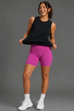 A woman modeling bright pink athletic biker shorts and a black sleeveless tank top, standing against a solid gray studio background. || Black