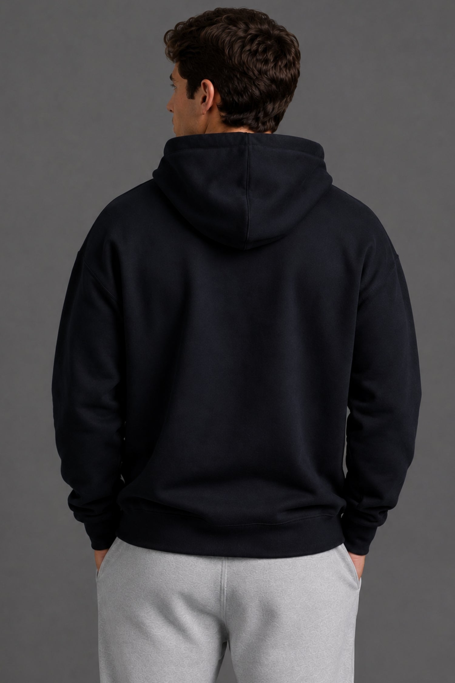 A man viewed from behind wearing a dark navy pullover hoodie and light gray sweatpants against a solid dark gray studio background.