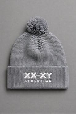 A light gray ribbed knit beanie with a matching pom-pom on top and a folded cuff featuring the white embroidered text 'XX-XY ATHLETICS', shown against a solid gray background. || Heather Gray