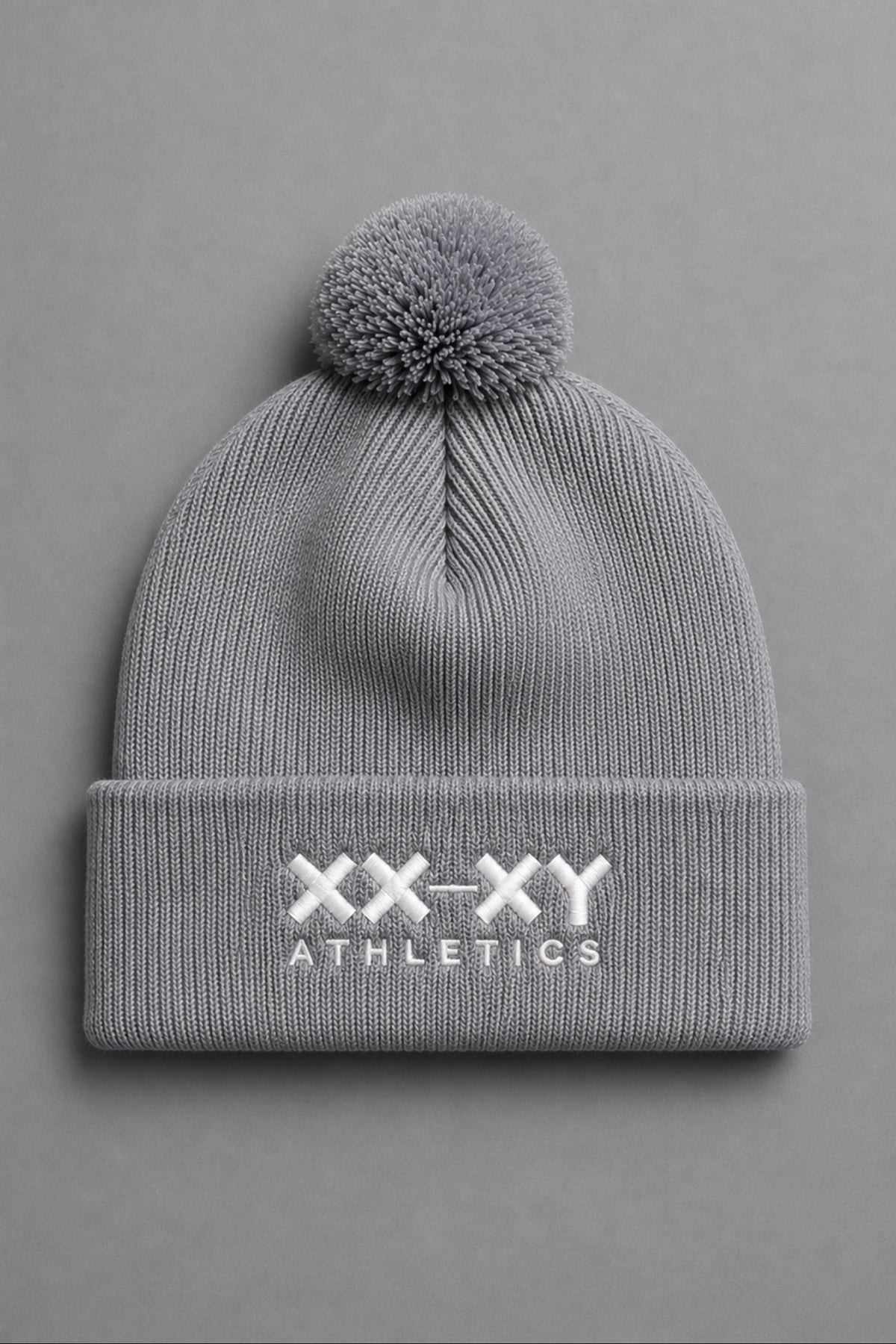 A light gray ribbed knit beanie with a matching pom-pom on top and a folded cuff featuring the white embroidered text 'XX-XY ATHLETICS', shown against a solid gray background. || Heather Gray