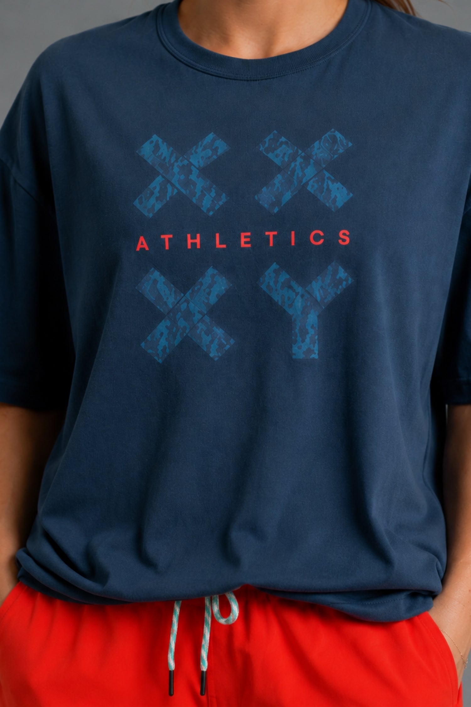 A person wearing a navy blue t-shirt with a blue distressed graphic of four 'X' shapes and the word 'ATHLETICS' in coral-red text, paired with red drawstring shorts.