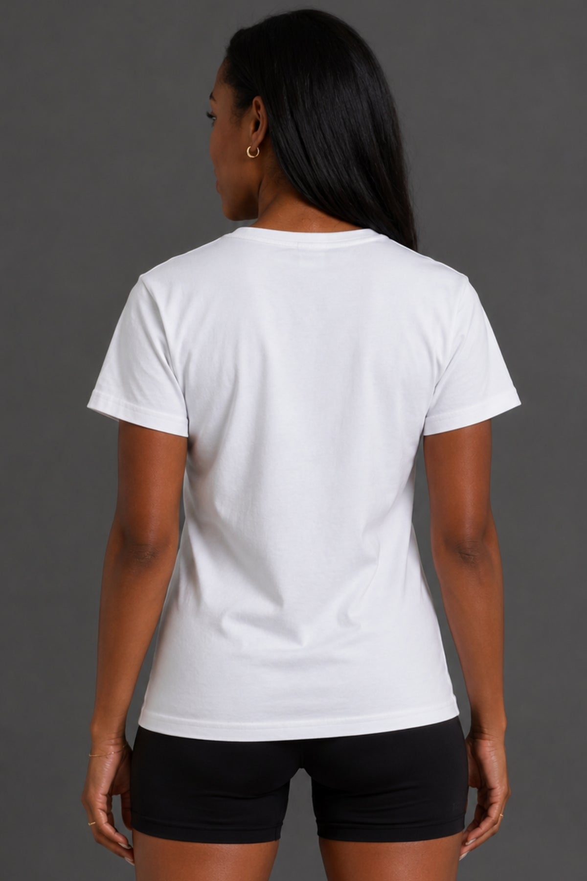 A woman viewed from the back wearing a plain white short-sleeve crew-neck T-shirt and black athletic shorts against a dark gray studio background. || White