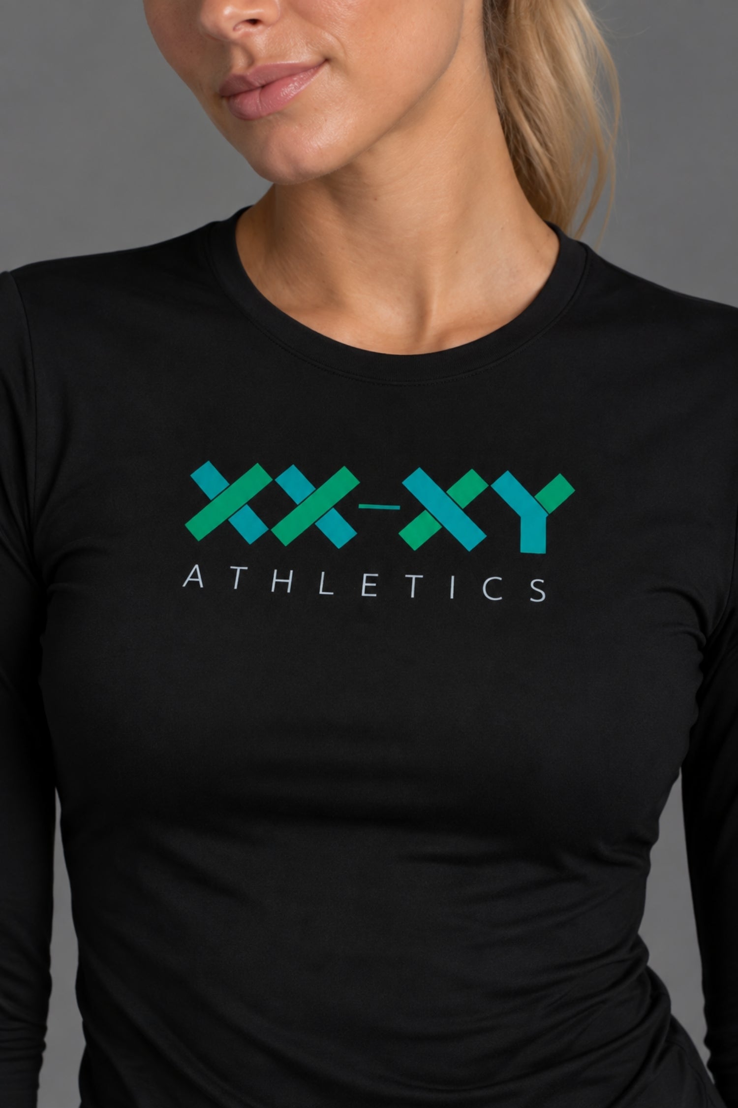 A close-up of a woman wearing a black long-sleeve athletic shirt with a teal and green 'XX-XY ATHLETICS' logo printed on the chest, set against a neutral gray background. || Black