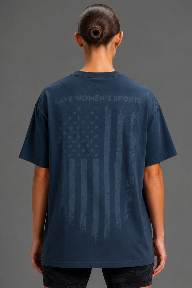 A woman viewed from the back wearing a navy blue t-shirt with a distressed American flag graphic and the words 'SAVE WOMEN'S SPORTS' printed across the upper back.