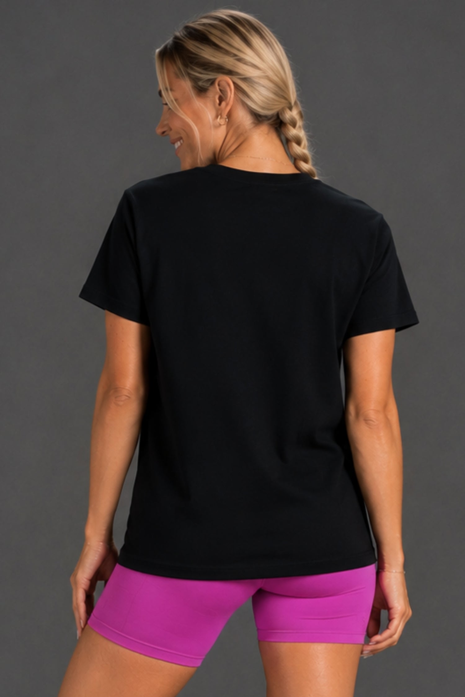 A woman shown from the back wearing a plain black short-sleeve t-shirt and bright pink athletic shorts against a dark gray studio background. || Bright Fuchsia