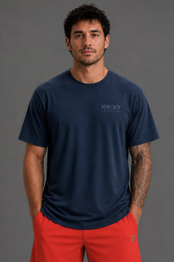 A man wearing a navy blue short-sleeve t-shirt with a subtle 'XX-XY athletik' logo on the left chest, paired with red athletic shorts, against a solid gray studio background. || Navy