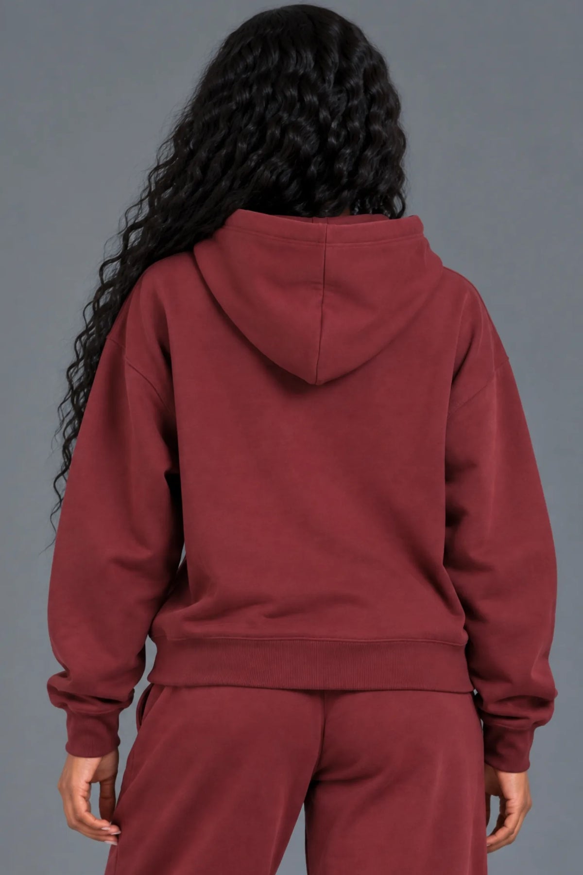 Back view of a woman wearing a blank burgundy pullover hoodie with matching sweatpants.