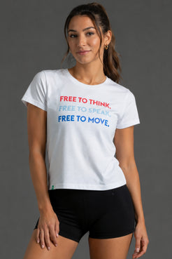 A woman modeling a white graphic t-shirt with the text 'FREE TO THINK, FREE TO SPEAK, FREE TO MOVE.' in red, light blue, and dark blue, paired with black athletic shorts against a dark gray studio background. || White