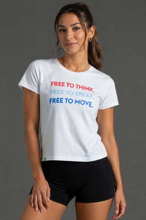 A woman modeling a white graphic t-shirt with the text 'FREE TO THINK, FREE TO SPEAK, FREE TO MOVE.' in red, light blue, and dark blue, paired with black athletic shorts against a dark gray studio background. || White