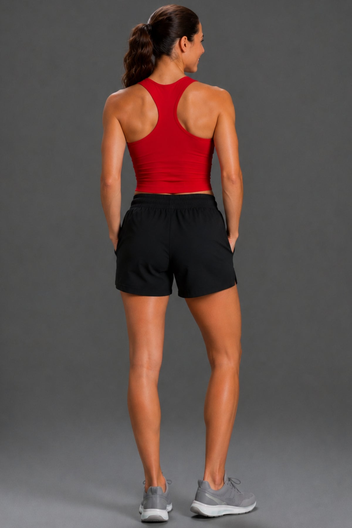 A woman shown from behind wearing a red athletic tank top and black athletic shorts, paired with gray sneakers, against a solid gray studio background.