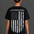 A woman viewed from the back wearing a black short-sleeve T-shirt with a distressed American flag graphic and the text 'SAVE WOMEN'S SPORTS' across the upper back, paired with white shorts against a dark gray studio background. || Black