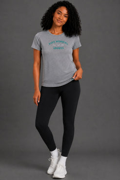 A woman modeling a heather gray t-shirt with 'SWEAT WOMEN'S SPORTS' printed in teal on the chest, paired with black leggings and white sneakers, against a solid gray studio background. || Heather Gray