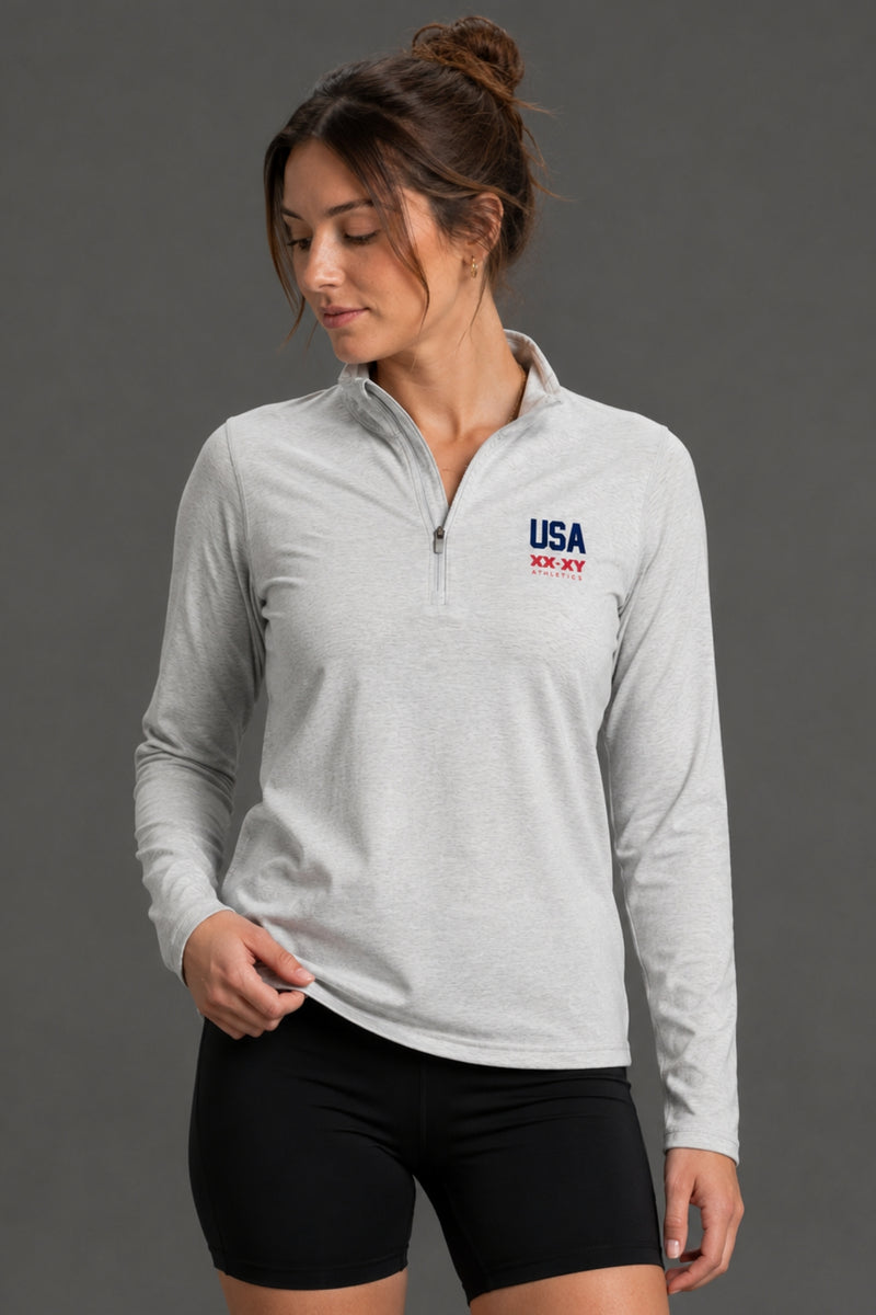 A woman modeling a light gray long-sleeve quarter-zip athletic pullover with a USA team logo on the left chest, paired with black athletic shorts and white sneakers, against a solid gray studio background. || Light Heather Gray - 1