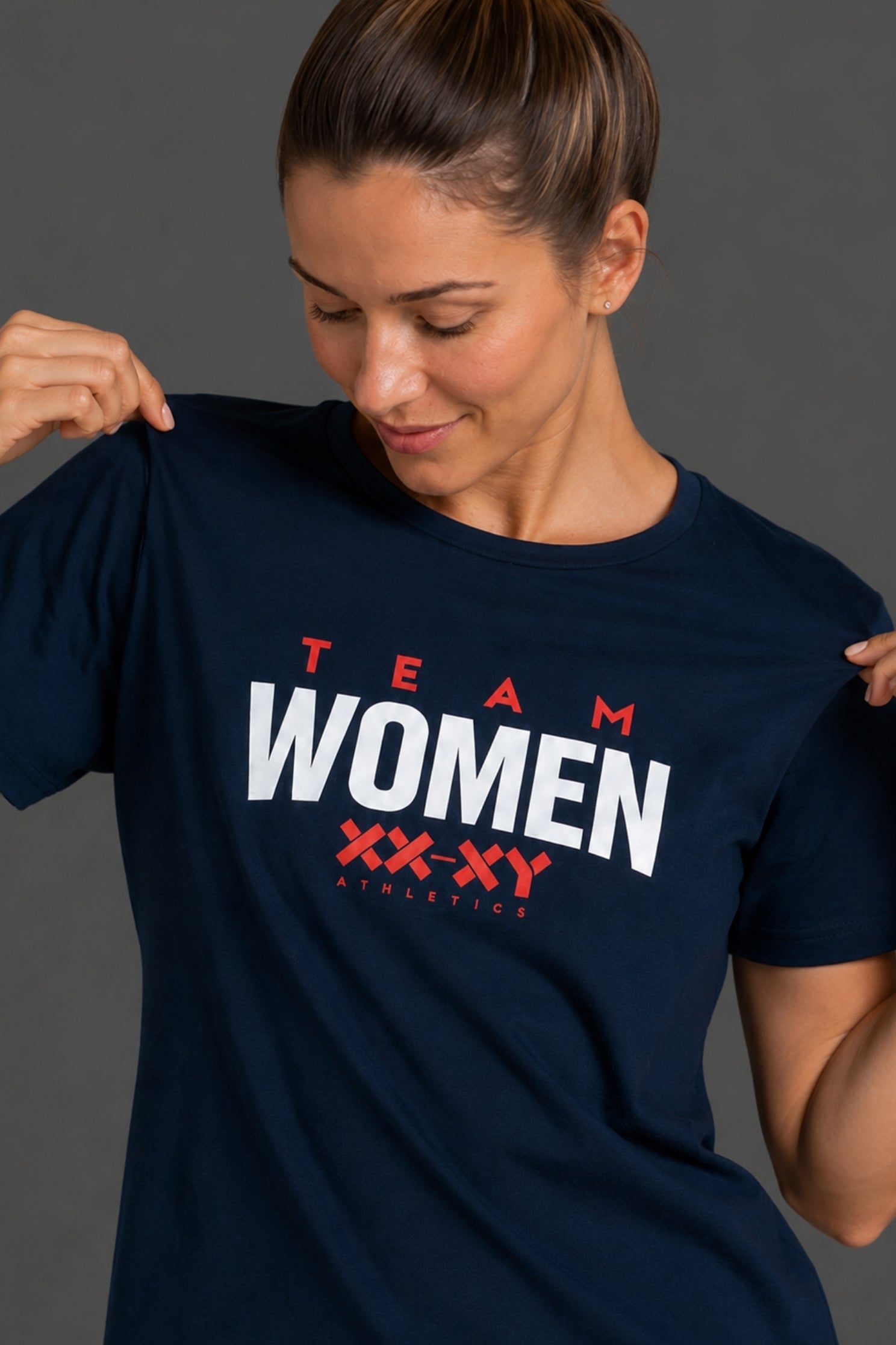A woman wearing a navy blue graphic t-shirt with 'TEAM WOMEN' printed in white and red on the chest, posing against a dark gray studio background. || Navy