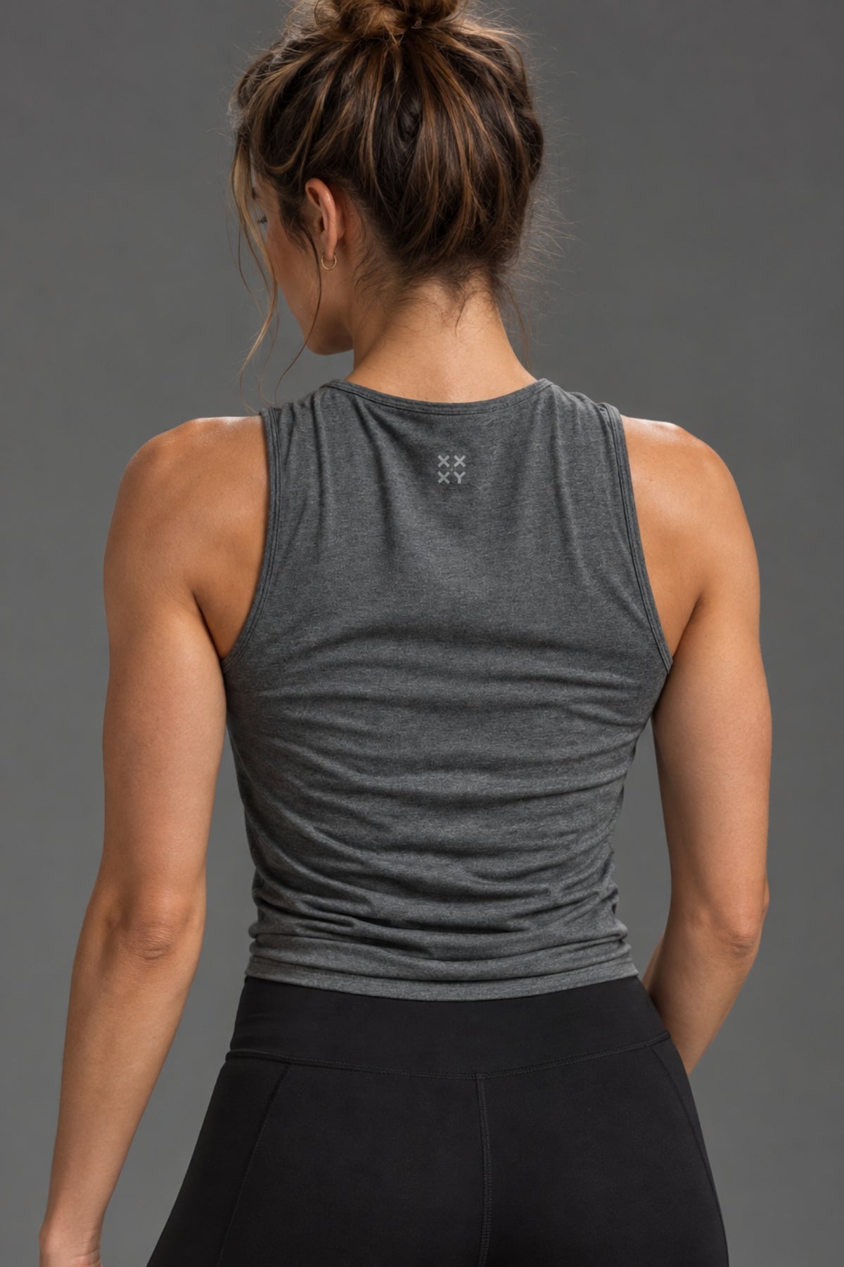 Rear view of a woman wearing a dark heather gray sleeveless athletic tank top and black leggings against a solid gray studio background.