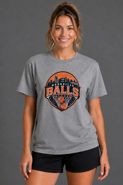 A woman wearing a heather gray t-shirt with a circular 'Nashville Balls' basketball graphic on the front, paired with black athletic shorts, against a solid gray studio background. || Heather Gray