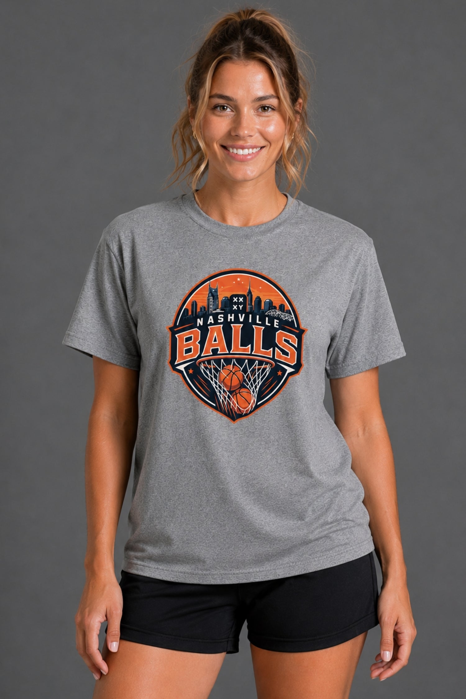 A woman wearing a heather gray t-shirt with a circular 'Nashville Balls' basketball graphic on the front, paired with black athletic shorts, against a solid gray studio background. || Heather Gray