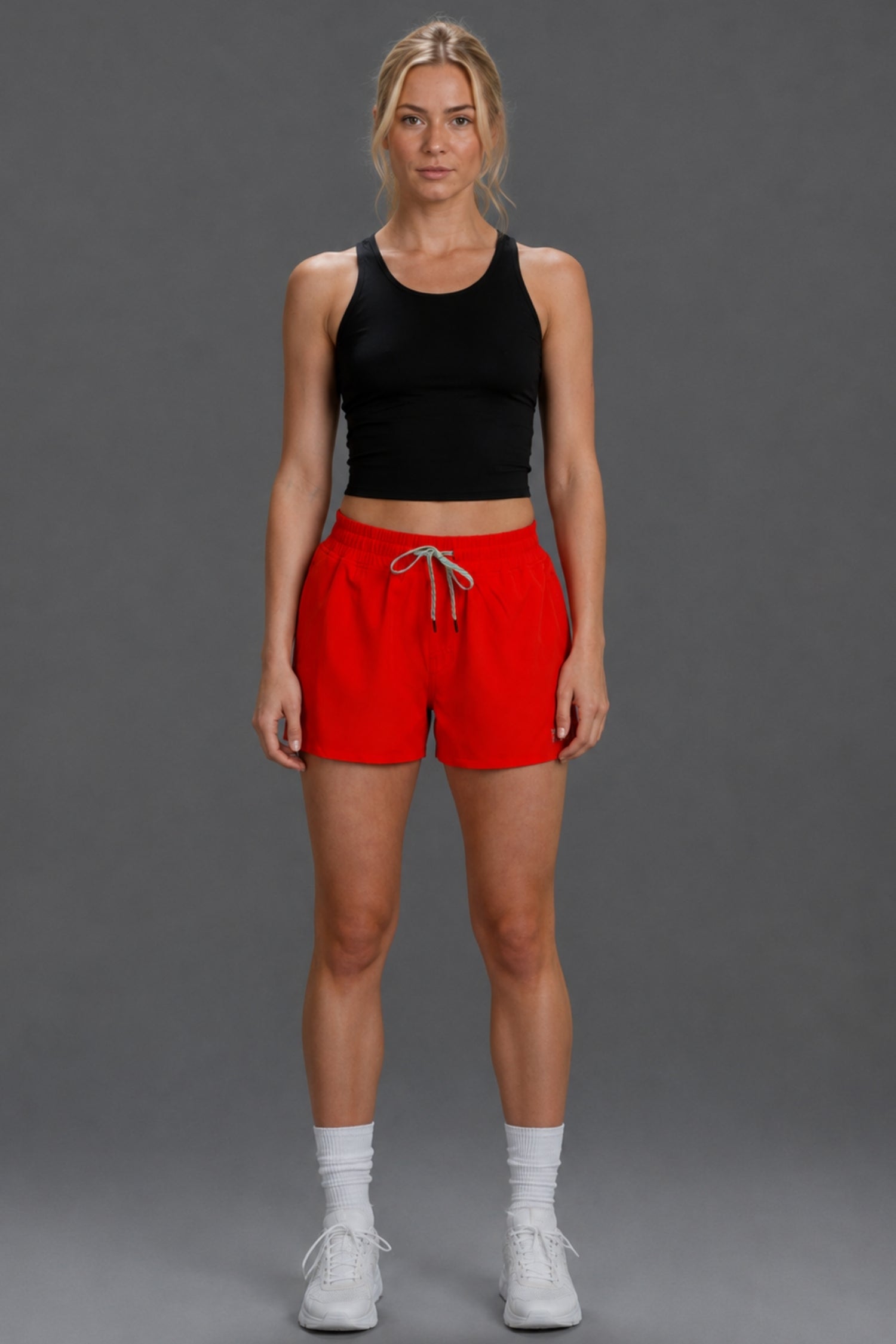 A woman stands against a plain gray studio background wearing a black sleeveless crop top and bright red athletic shorts with a white drawstring, paired with white socks and white sneakers. || Red