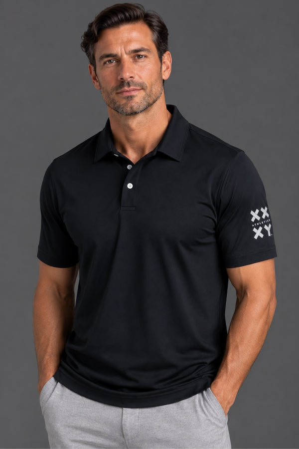 A man modeling a dark navy polo shirt with white embroidered logos on the sleeve, paired with light gray jogger pants, against a solid gray studio background. || Black