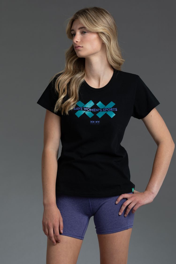 Woman wearing a black t-shirt with a logo and purple shorts on a gray background || Teal