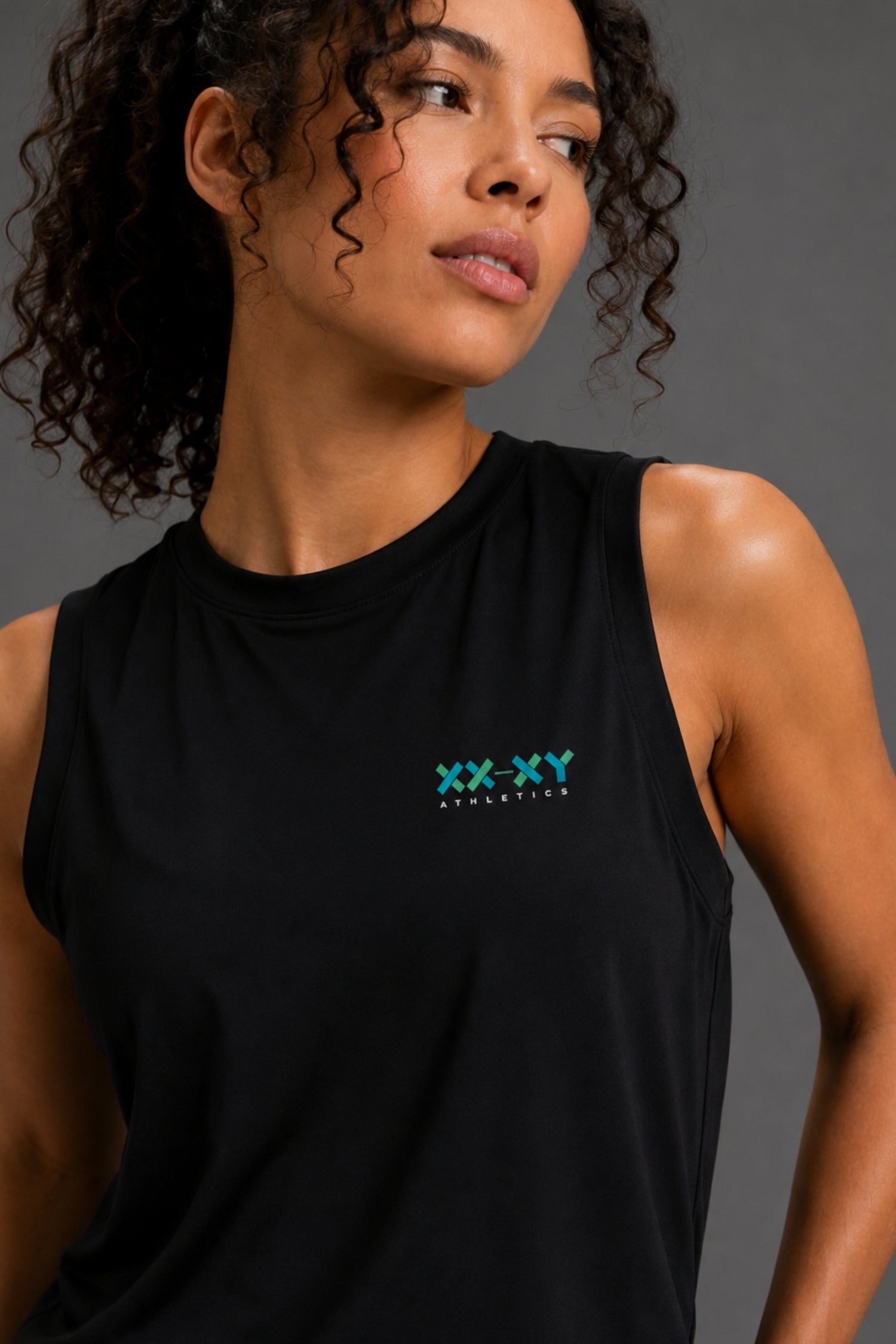 A close-up of a woman wearing a black sleeveless tank top with a small green and blue 'XX-XY STATIONERY' logo on the left chest, set against a dark gray studio background. || Black