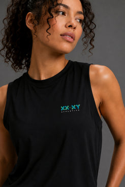 A close-up of a woman wearing a black sleeveless tank top with a small green and blue 'XX-XY STATIONERY' logo on the left chest, set against a dark gray studio background. || Black