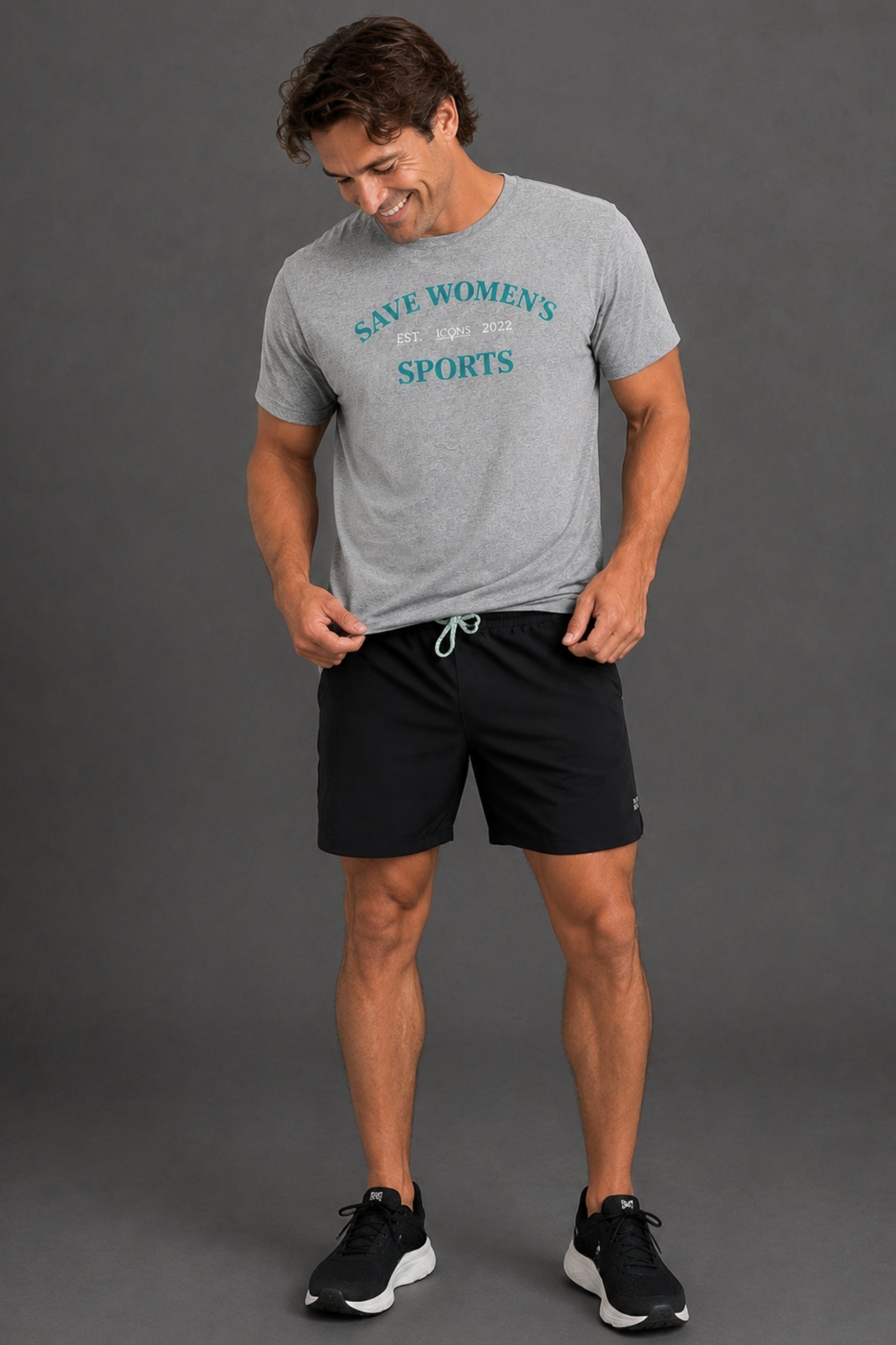 A man wearing a heather gray graphic t-shirt with 'SAVE WOMEN'S SPORTS' in teal text, paired with black athletic shorts and black sneakers, standing against a solid gray studio background. || Heather Gray