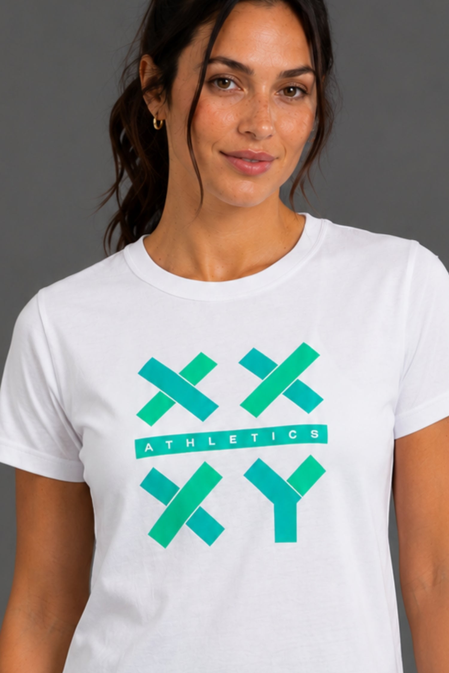 A woman wearing a white crewneck t-shirt with a teal and green geometric 'XX ATHLETICS YY' graphic on the front, set against a solid gray studio background. || White