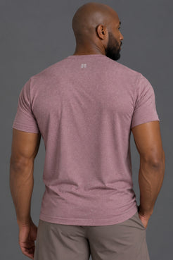 A man viewed from the back wearing a heathered dusty rose short-sleeve t-shirt and taupe pants, set against a solid dark gray studio background. || Burgundy Heather